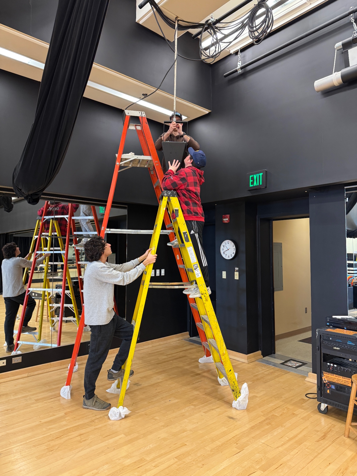 A team installing an overhead loudspeaker from two tall step ladders in a dance studio.