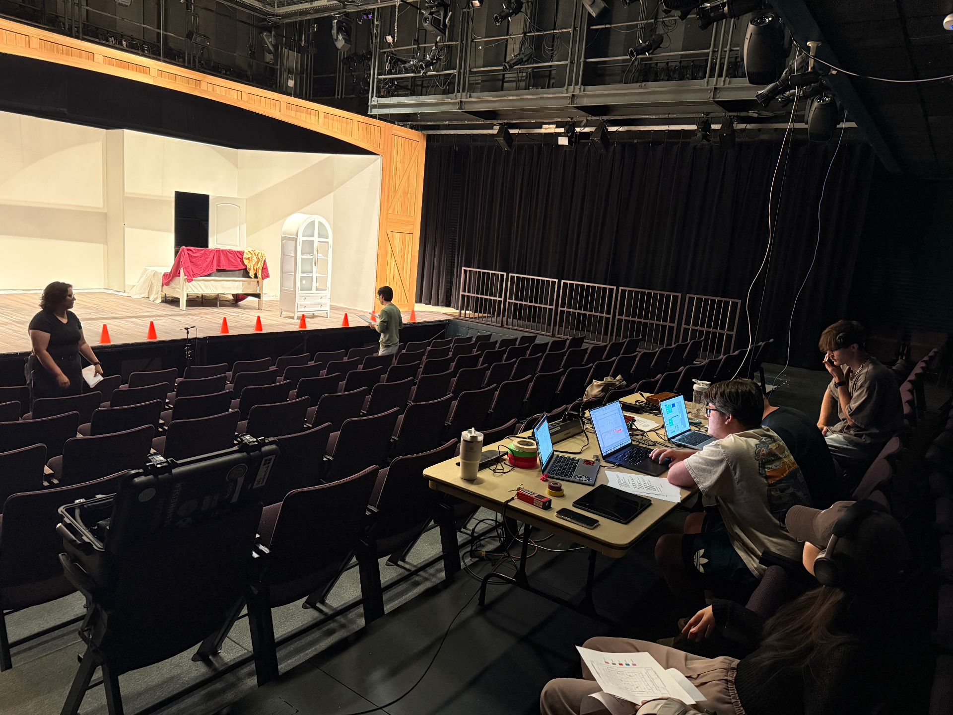 Tech rehearsal with students at a production table in the house of a thrust theatre, set with a small bedroom interior on the stage.